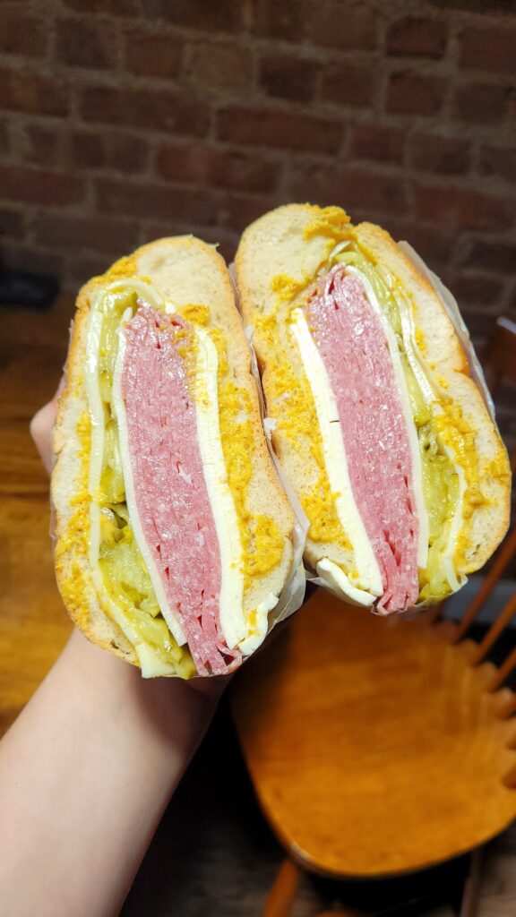 A hand holding a sub sandwich cut in half, showing layers of salami, cheese, pickles, mustard, and other condiments.